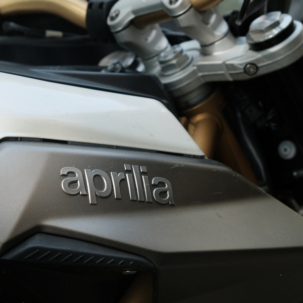 Close-up of an Aprilia motorcycle tank with the logo prominently displayed, featuring a sleek metallic design and parts of the handlebar visible